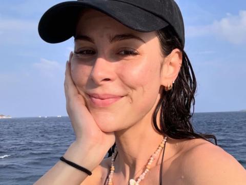Lena Meyer-Landrut sitzt lächelnd mit Cap und Bikini am Meer