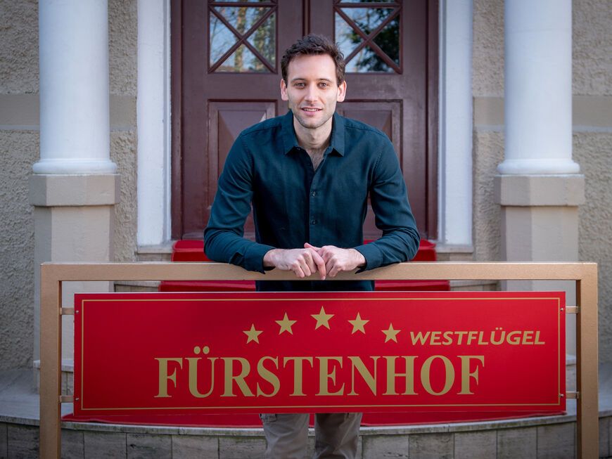 Sturm der Liebe Julian Specht lehnt an einem Fürstenhof-Schild vor dem Hotel