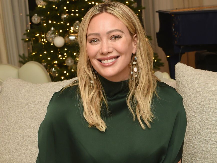 Hilary Duff sitzt lächelnd auf der Couch, im Hintergrund steht ein Weihnachtsbaum