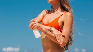 Frau im Bikini am Strand cremt sich mit Sonnencreme ein vor blauem Himmel