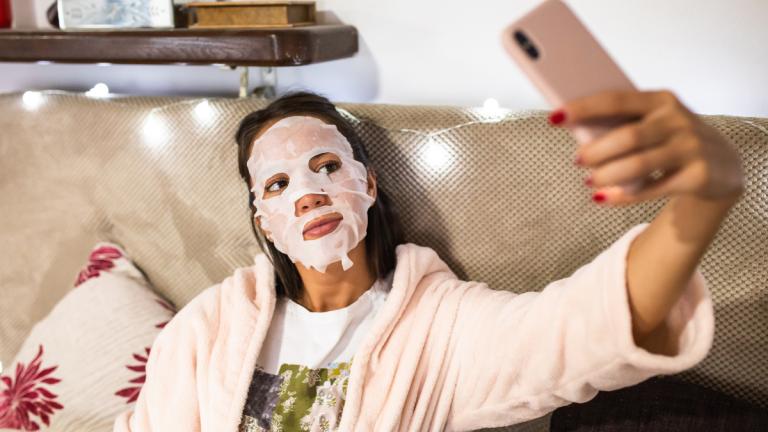 Frau sitzt mit Tuchmaske auf Bett und macht Selfie