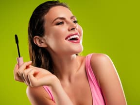 Frau mit Mascara steht lachend vor grüner Wand