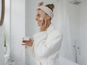 Junge Frau lächelt in Spiegel und benutzt Anti-Aging-Creme