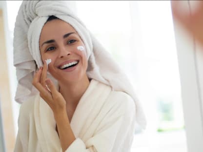 Anti-Aging-Creme ab 30 Frau cremt sich lächelnd vor Spiegel ein