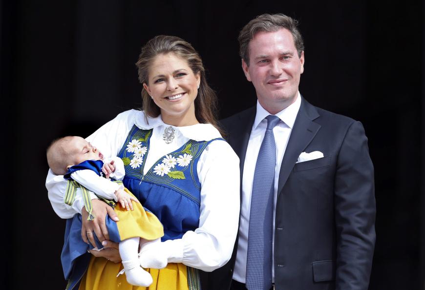 Am schwedischen Nationalfeiertag tritt Prinzessin Madeleine mit Baby Leonore im Partner-Trachten-Look vor die Presse. Chris O'Neill bleibt bei einem dunkelblauen Anzug.