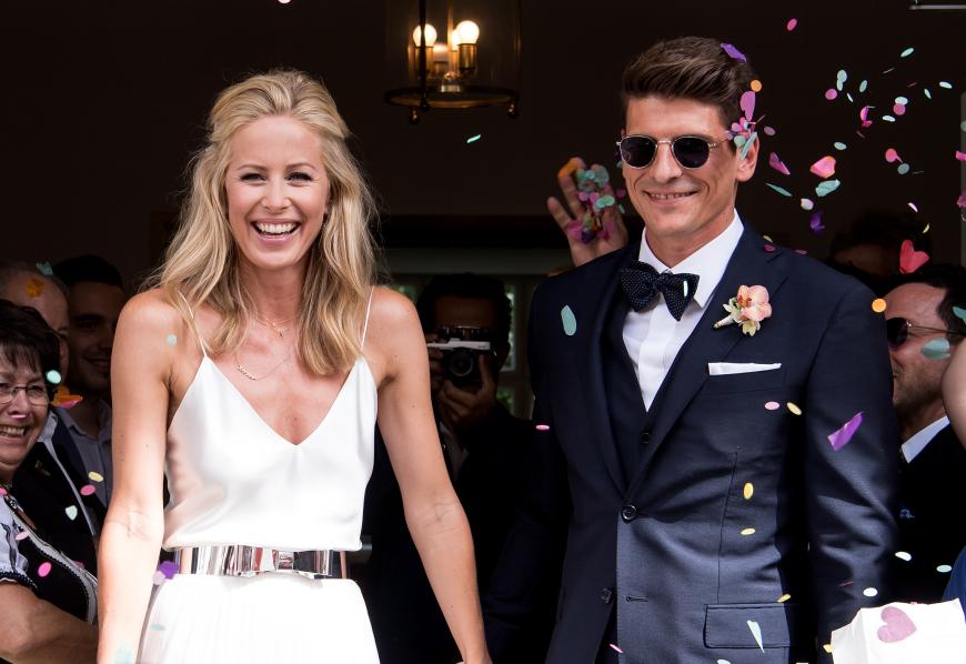 Mann und Frau: Mario Gomez mit lässiger Sonnenbrille und Fliege und Carina in einem schlichten Traum aus Weiß mit süßem Schleifengürtel.