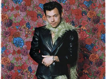 Harry Styles mit Federboa und schwarzem Anzug vor einer Blumenwand