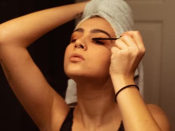 Frau mit Handtuch auf dem Kopf trägt Mascara vor Spiegel auf