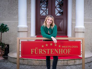 Greta Bergmann steht hinter dem Fürstenhof-Schild und lacht