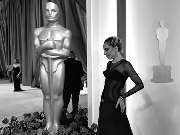 Schwarz-weiß Foto von Lady Gaga auf dem Oscars-Teppich die nach unten guckt neben einer Oscars-Statue