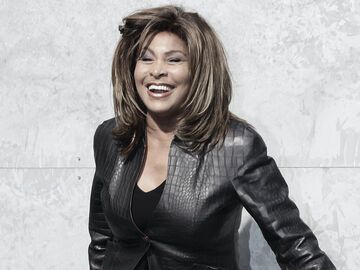Tina Turner steht lachend vor einer grauen Wand