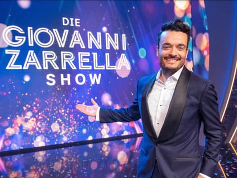 Giovanni Zarrella grinst und zeigt mit ausgestrecktem Arm auf eine Leinwand mit dem Logo der Giovanni Zarrella Show