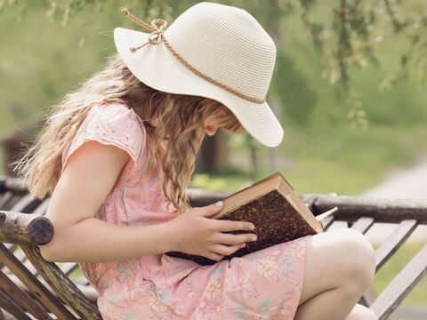 Mädchen liest Buch im Garten und trägt einen Hut und ein Kleid.