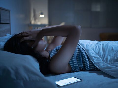 Frau liegt mit Smartphone im Bett, greift sich an den Kopf