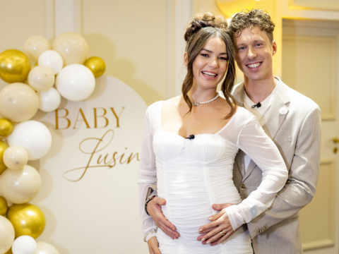 Valentin Lusin steht hinter Renata Lusin in einem weißen Kleid und umarmt sie bei ihrer Babyparty