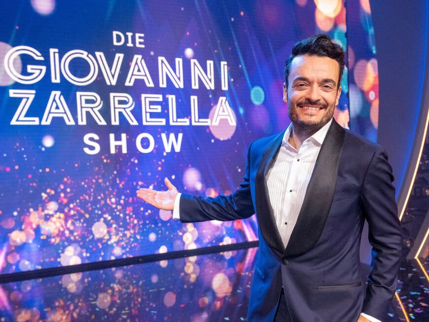 Giovanni Zarrella grinst und zeigt mit ausgestrecktem Arm auf eine Leinwand mit dem Logo der Giovanni Zarrella Show