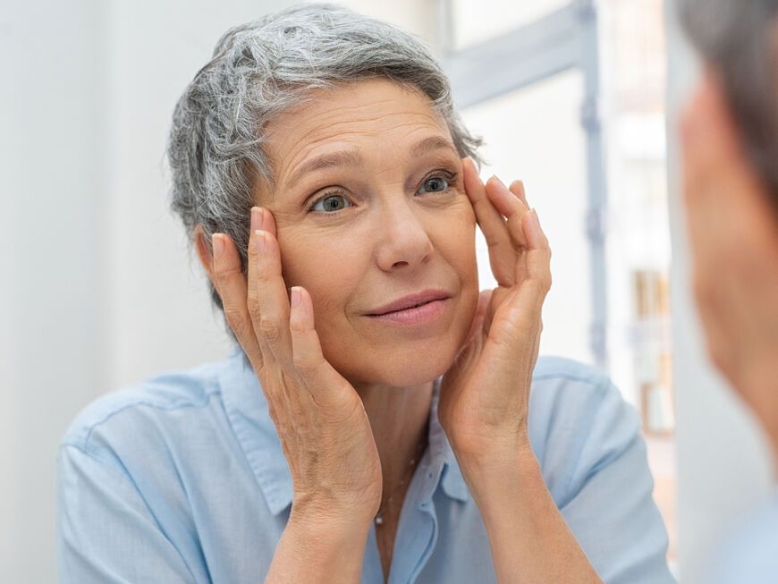 Ältere Frau mit kurzen, grauen Haaren betrachtet Falten im Spiegel