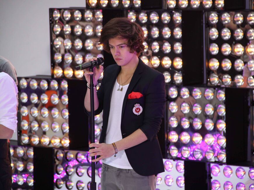 Harry Styles steht auf der Bühne und guckt ernst mit der Hand am Mikro 2012