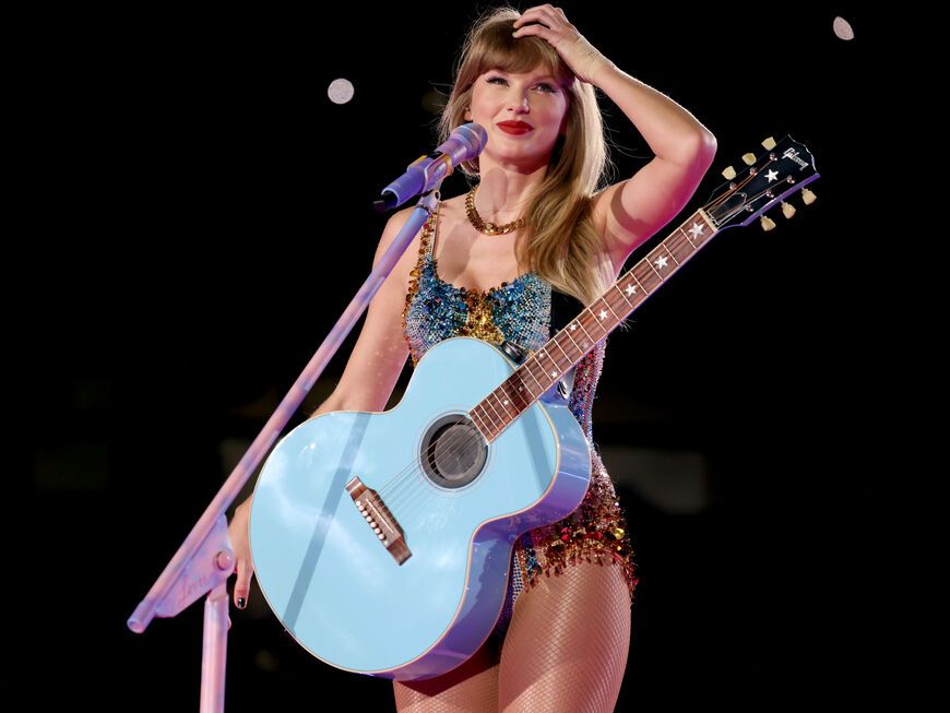 Taylor Swift mit Gitarre um den Bauch lächelt und fährt sich durch die Haare