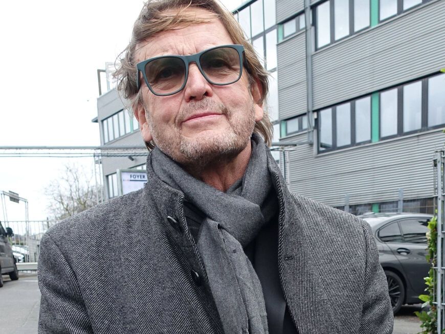 Peter Althof schaut ernst mit Sonnenbrille und Schal vor Gebäude