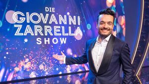 Giovanni Zarrella grinst und zeigt mit ausgestrecktem Arm auf eine Leinwand mit dem Logo der Giovanni Zarrella Show