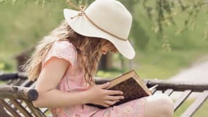 Mädchen liest Buch im Garten und trägt einen Hut und ein Kleid.