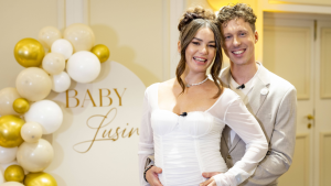 Valentin Lusin steht hinter Renata Lusin in einem weißen Kleid und umarmt sie bei ihrer Babyparty
