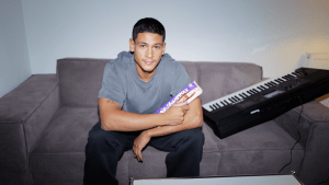 Emilio Sakraya sitzt auf einer Couch mit einer Tafel Milka in der Hand und einem Keyboard neben sich