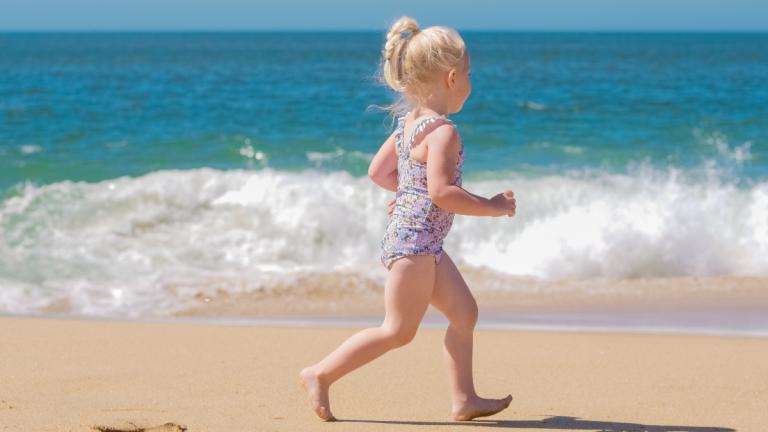 Blondes Mädchen rennt am Strand vor dem Meer rum und trägt einen Badeanzug