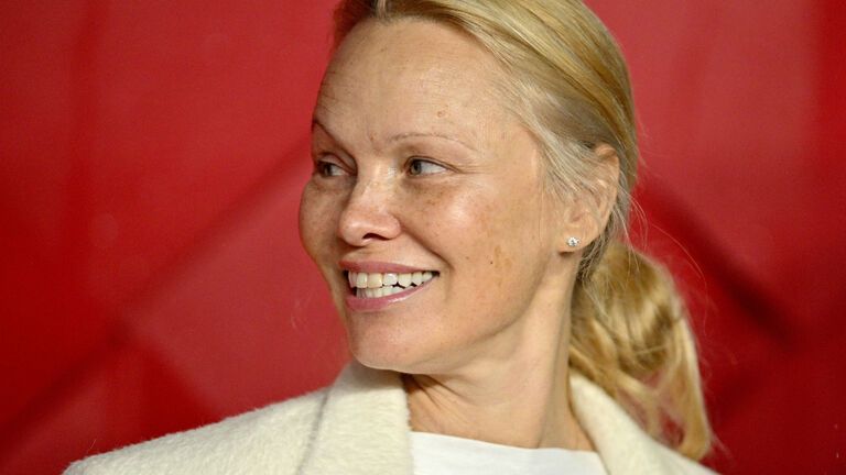 Pamela Anderson guckt zur seite und lächelt vor rotem Hintergrund